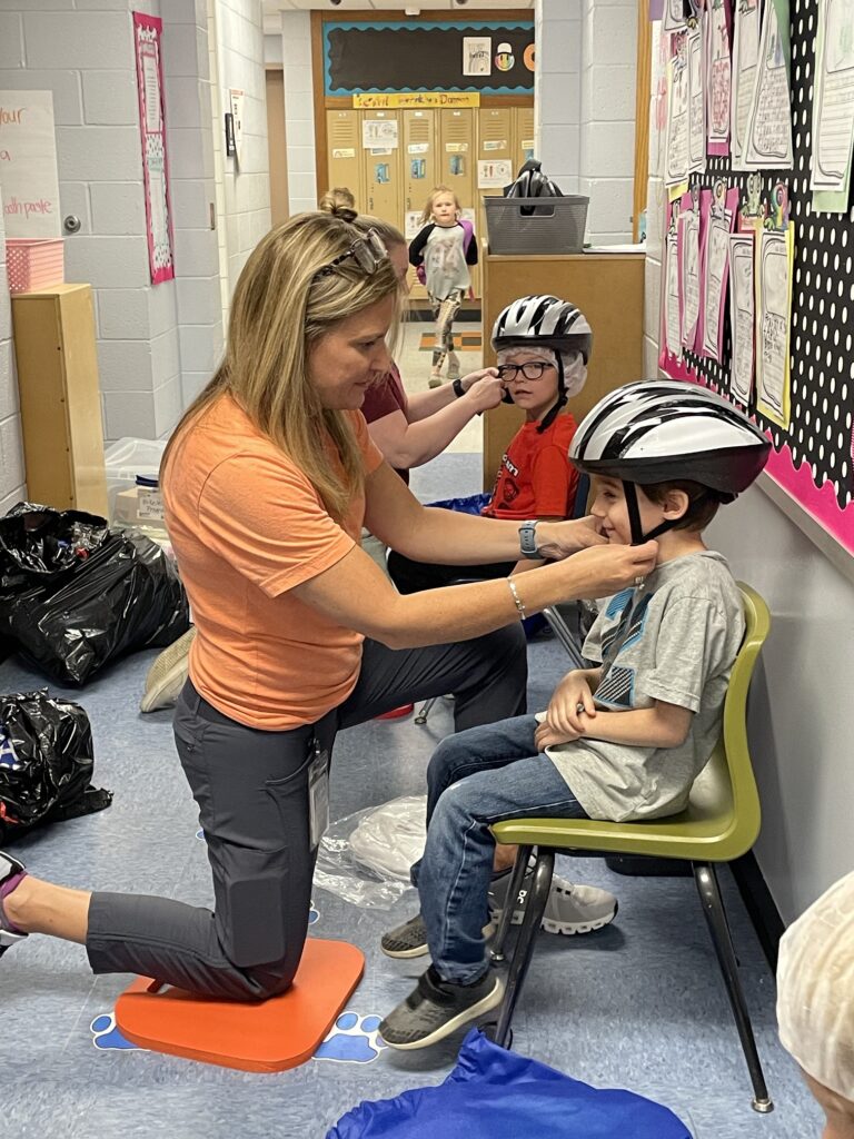 Picture shows a white woman kneeling in front of a sitting child to properly fit a helmet on the child's head