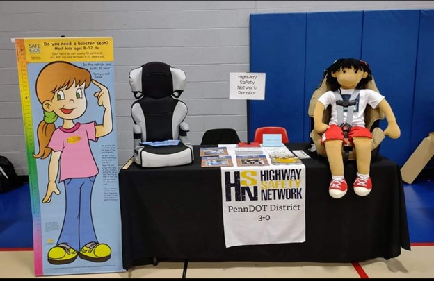 Picture shows a growth chart showing how tall a child needs to be before moving out of a booster seat next to a table with two child passenger safety seats on display with other paper resources between them