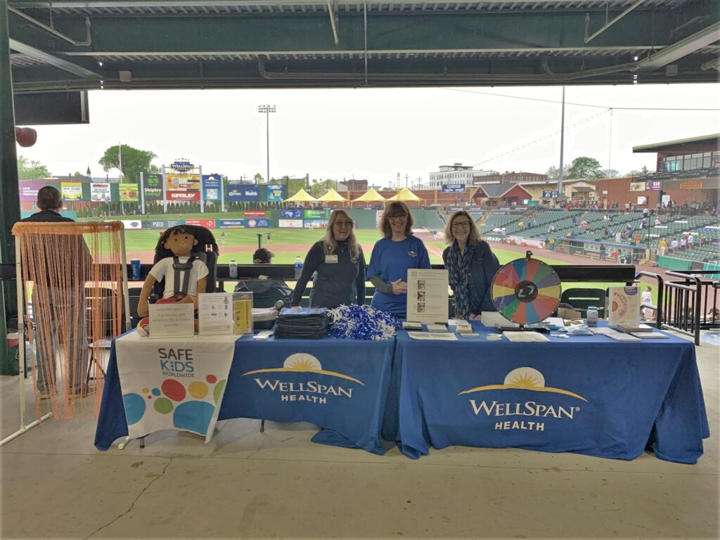 Picture shows three white women smiling behind two tables with educational materials and displays on child passenger safety at a community event