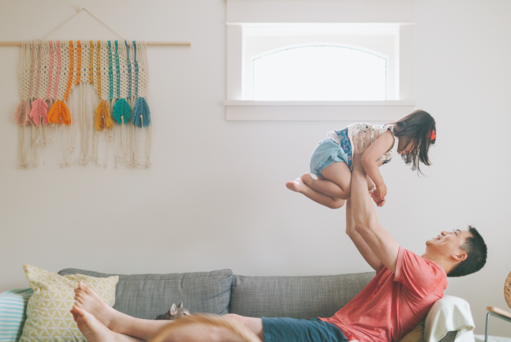 Father laying back on a couch, lifting his daughter up above his head